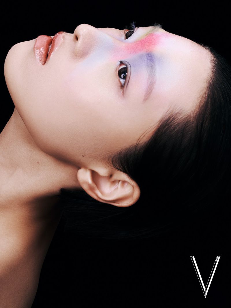 Close-up studio portrait of a young woman with colorful makeup, facing away from the camera against a dark background. 'V' logo at the bottom.