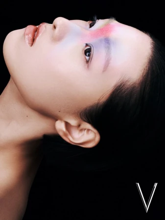 Close-up studio portrait of a young woman with colorful makeup, facing away from the camera against a dark background. 'V' logo at the bottom.