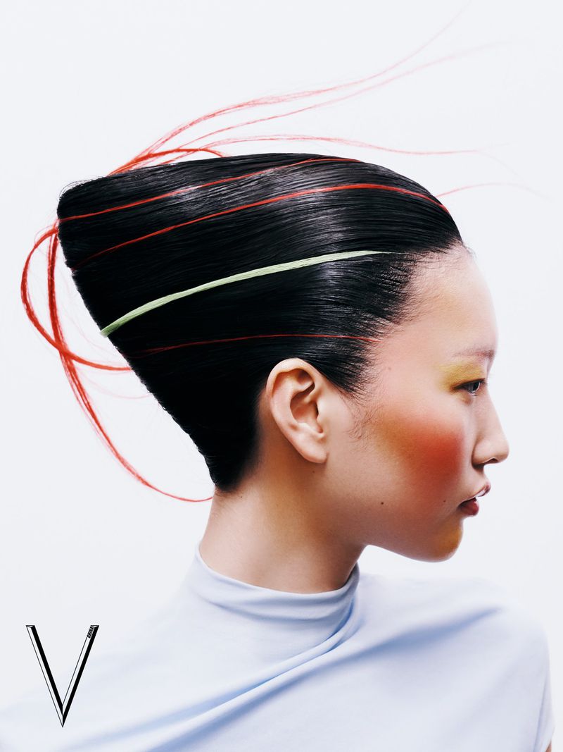 Portrait of an Asian woman with an elegant hairstyle and stylized makeup in a light blue top, set against a bright white backdrop.