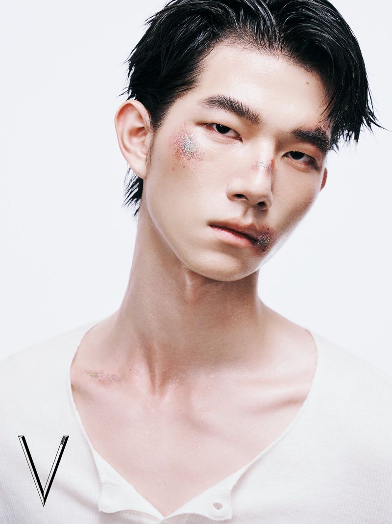 Portrait of a dark-haired Asian male model with light skin and glittery makeup posing in a photo studio with a white backdrop and a white shirt.