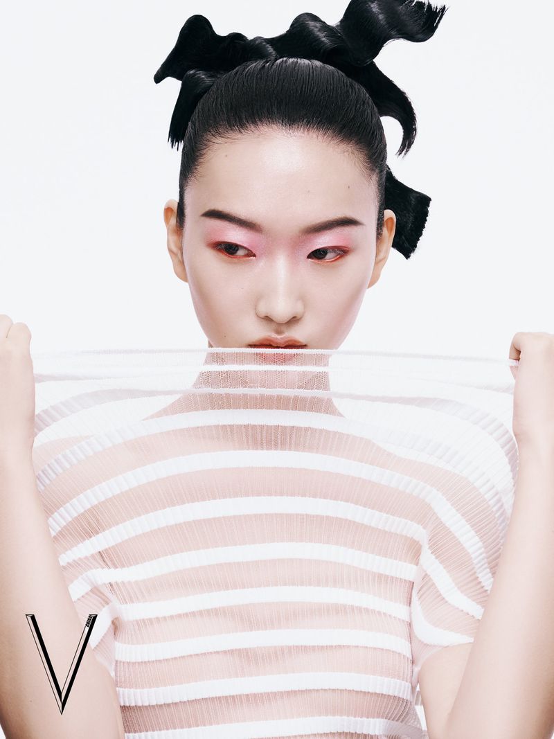 Asian woman poses with dramatic eye makeup and black hair in a studio portrait, wearing a striped transparent blouse, with logo in the corner.