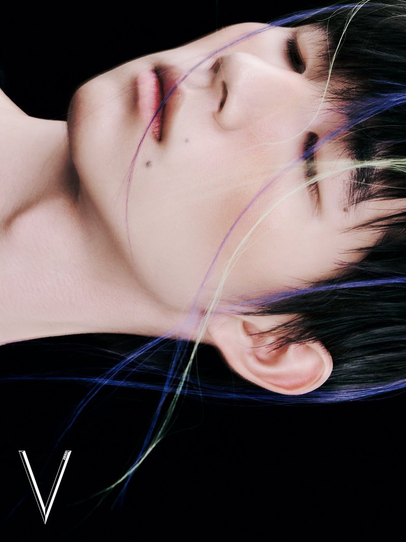 Close-up studio shot of a person with styled black hair with colorful strands, eyes closed, against a black background and 'V' text.