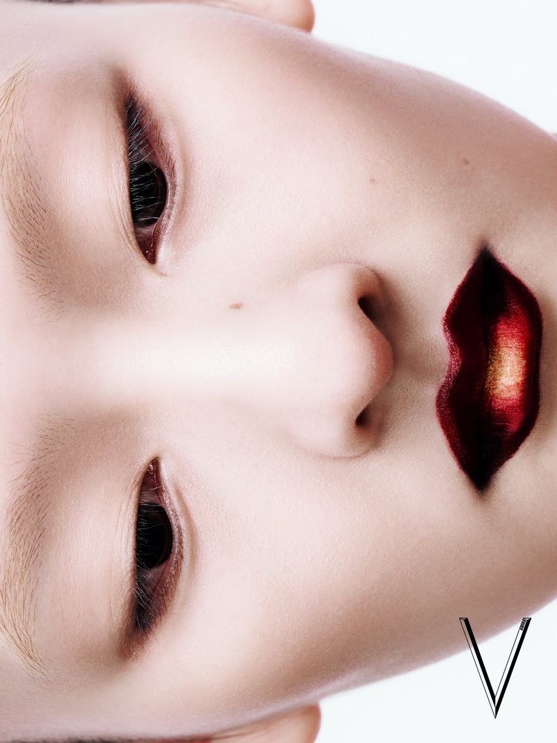 Dramatic close-up portrait of a young adult female with intense makeup and dark red lipstick, set against a stark white background.