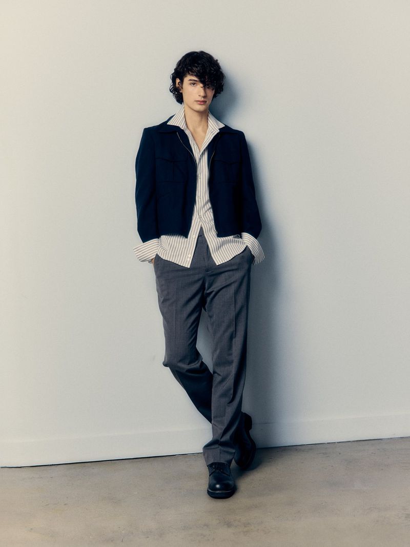 Young adult male in a navy cardigan, striped shirt, and gray pants leans against a light gray wall in a studio setting.