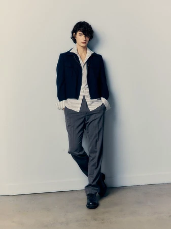 Young adult male in a navy cardigan, striped shirt, and gray pants leans against a light gray wall in a studio setting.