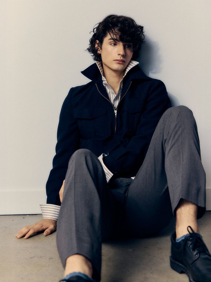 Male fashion model in a dark blue jacket and grey trousers seated indoors against a wall in a casual pose, glancing to the side.