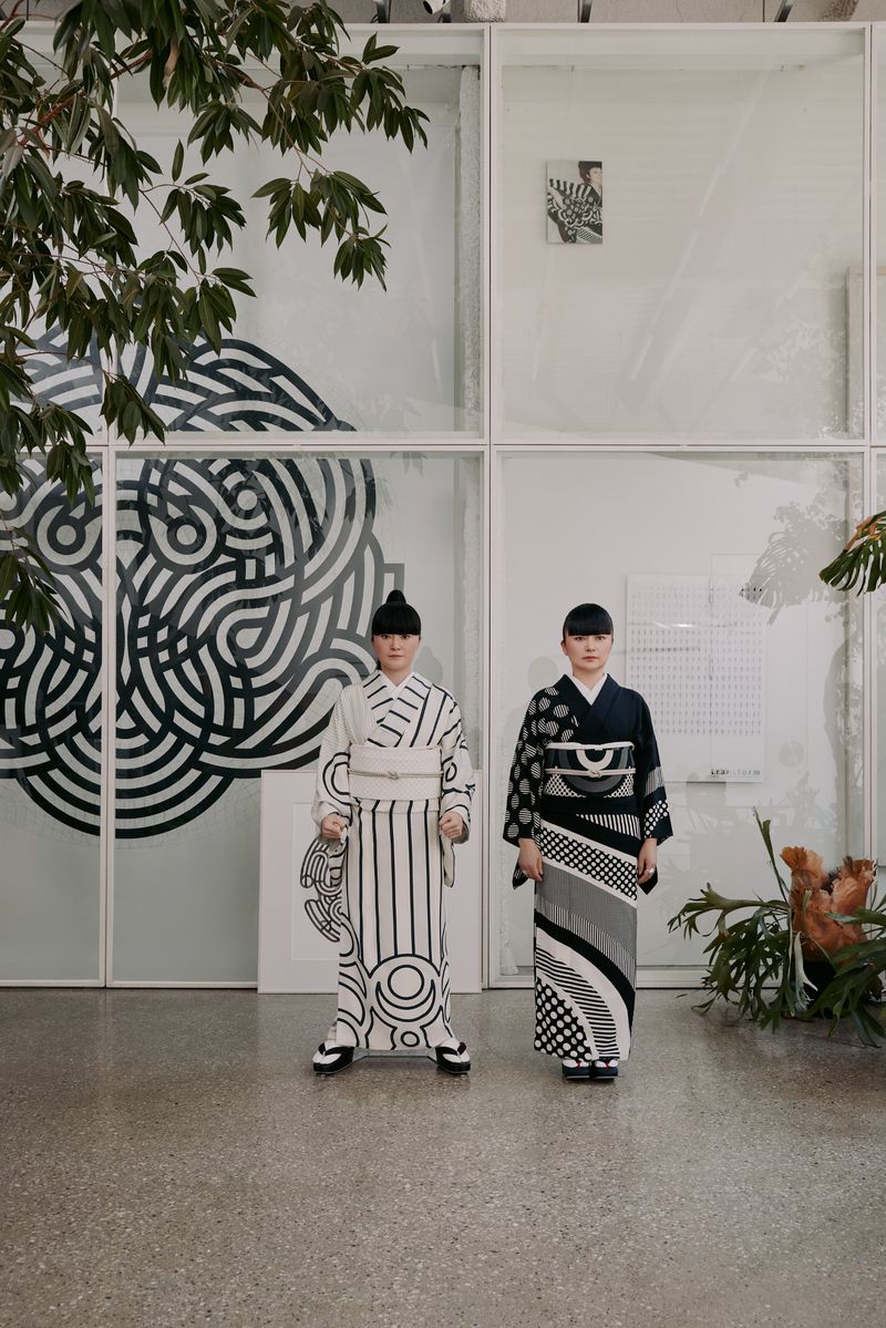 Two women in patterned kimonos stand in front of a white-framed glass wall with black circular designs and green plants, fashion photoshoot.