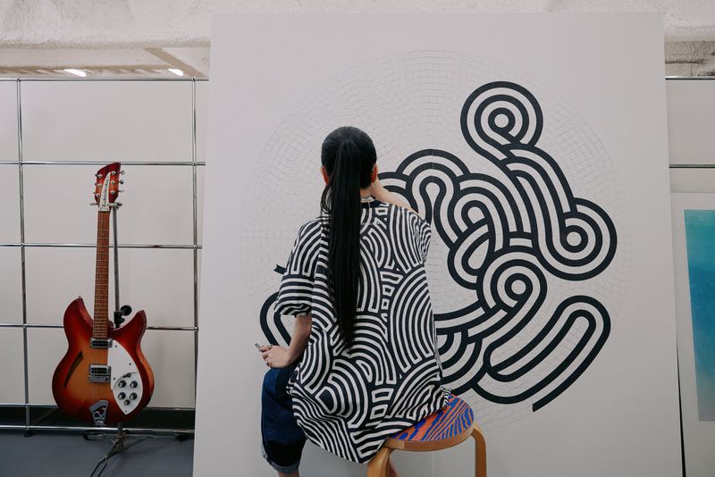 Back view of an adult female artist creating black and white line art on a canvas, seated on a stool in a brightly lit indoor space.