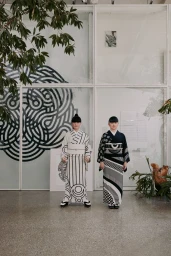 Two women in matching patterned kimonos standing indoors in a bright gallery space with graphic wall art and plants.