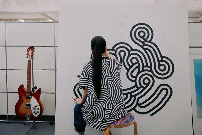 Woman seen from behind wearing a black and white patterned shirt, seated on a stool and painting a large canvas.