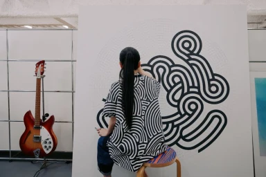 Woman seen from behind wearing a black and white patterned shirt, seated on a stool and painting a large canvas.