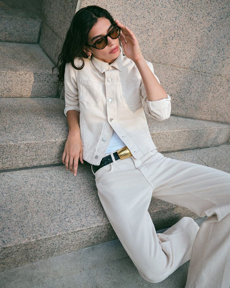 A woman with sunglasses poses while wearing a beige jacket, pants, and a gold belt while sitting on concrete stairs in an urban setting.