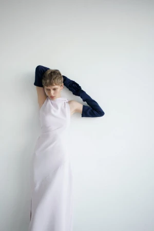 Fashion shot of a young adult woman in a lilac dress and navy gloves posing against a white wall, looking down in a studio setting.