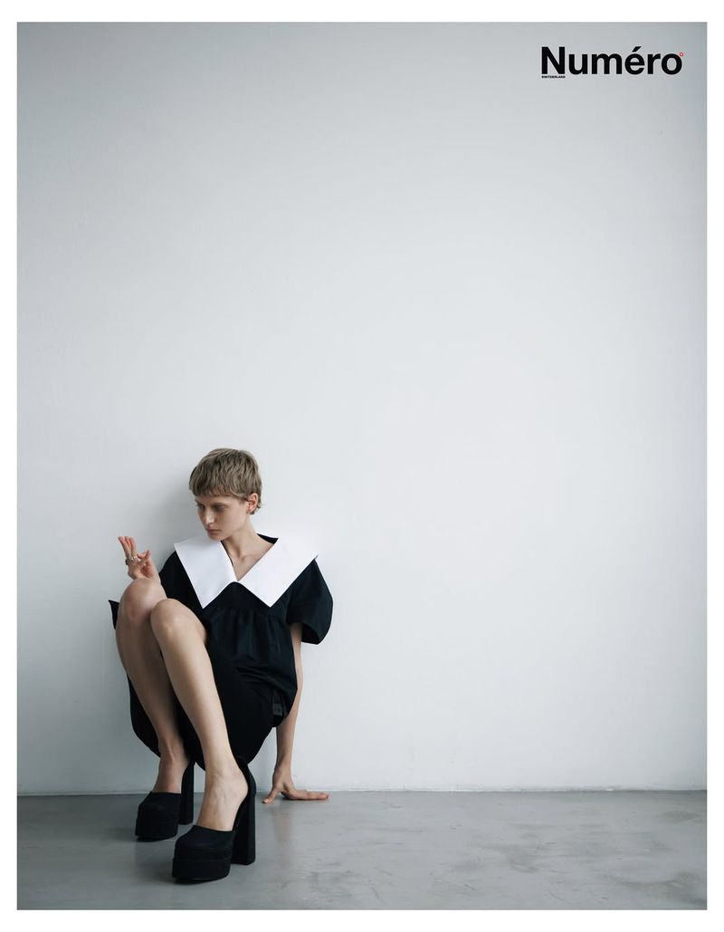 Fashion editorial shot of a woman in a black dress with a large white collar and platform heels, seated against a white wall with magazine logo.
