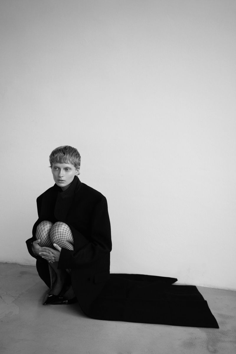 A person with short hair crouches on the floor wearing fishnet stockings and an oversized black coat against a white wall backdrop.