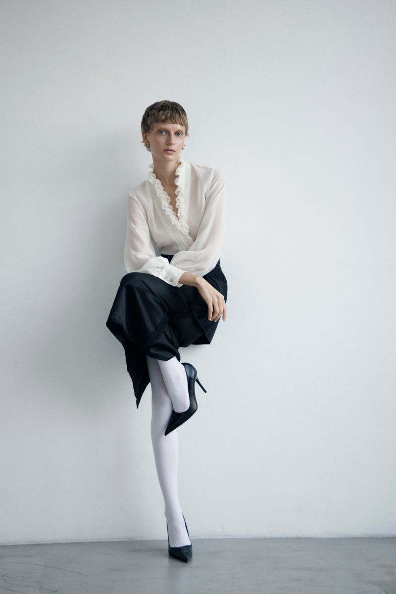 Full-length studio portrait of a slender woman with short hair, wearing a white blouse, black skirt, white tights and black heels, leaning against a wall.
