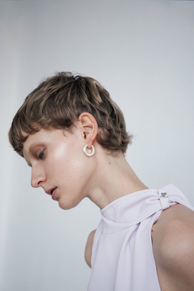 Profile view of a fair-skinned adult woman with short brown hair wearing a light purple top and silver hoop earrings, against a white background.