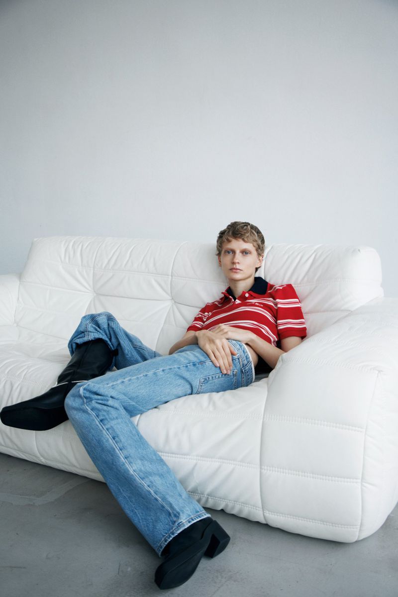 A person lounges on a white couch wearing jeans, a red-and-white striped collared shirt, and black boots against a plain white backdrop.