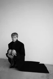 Model wearing a dark coat and fishnet tights squatting against a plain white studio background in monochrome.