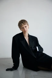 Model with short hair wearing an oversized black blazer and black leather gloves seated against a white background