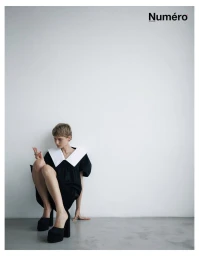 Model with short hair wearing a black dress with a large white collar and platform heels against a light background