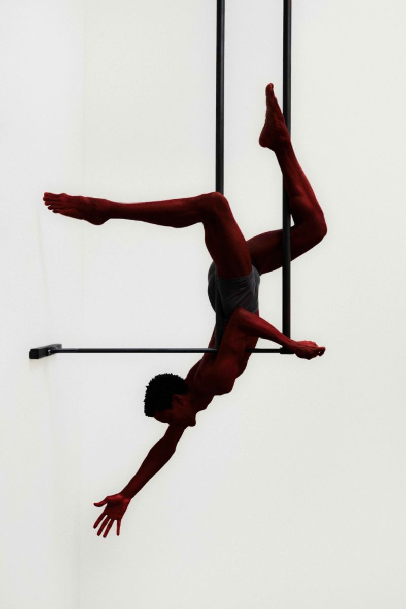 A male acrobat performs a dynamic aerial pose on a metal trapeze against a stark white backdrop in a studio setting.
