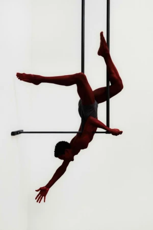 A male acrobat performs a dynamic aerial pose on a metal trapeze against a stark white backdrop in a studio setting.