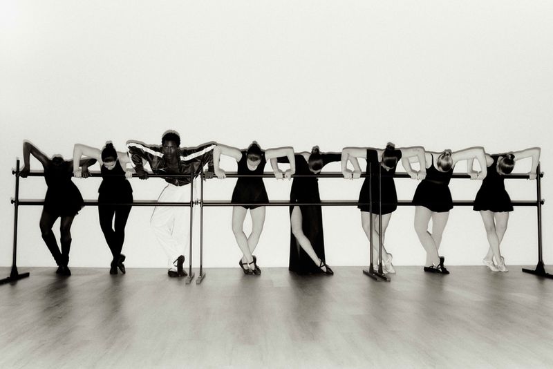Eight ballet dancers in various poses at a barre in a dance studio, lit by diffused light against a white backdrop. Focus on form and movement.