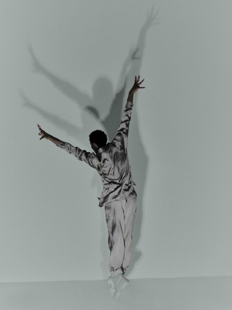 A person in a silky outfit is captured mid-jump in a studio, arms and legs outstretched, casting a large shadow against a light blue backdrop.