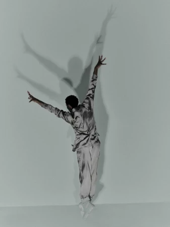 A person in a silky outfit is captured mid-jump in a studio, arms and legs outstretched, casting a large shadow against a light blue backdrop.