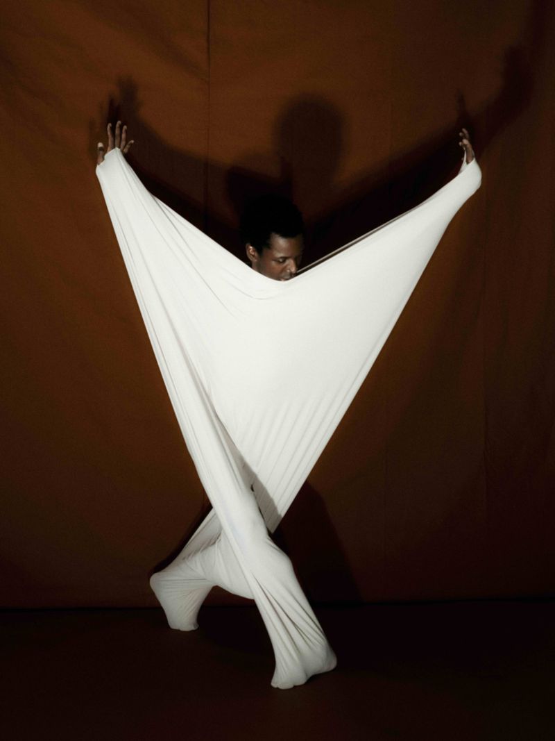 Portrait of a Black man with expressive pose and white draped clothing against brown background in studio environment with soft lighting.