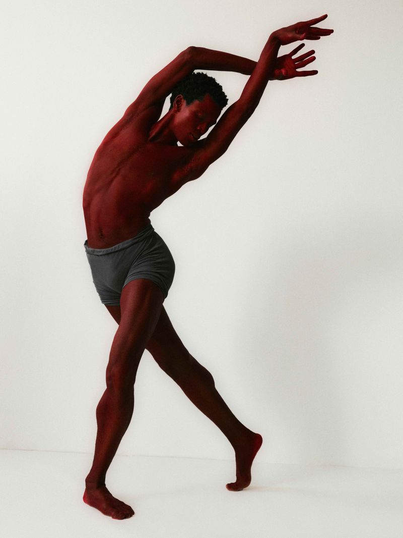 An adult male is posing in a dance-like stance with arms arched overhead against a white backdrop in a studio or simple setting.