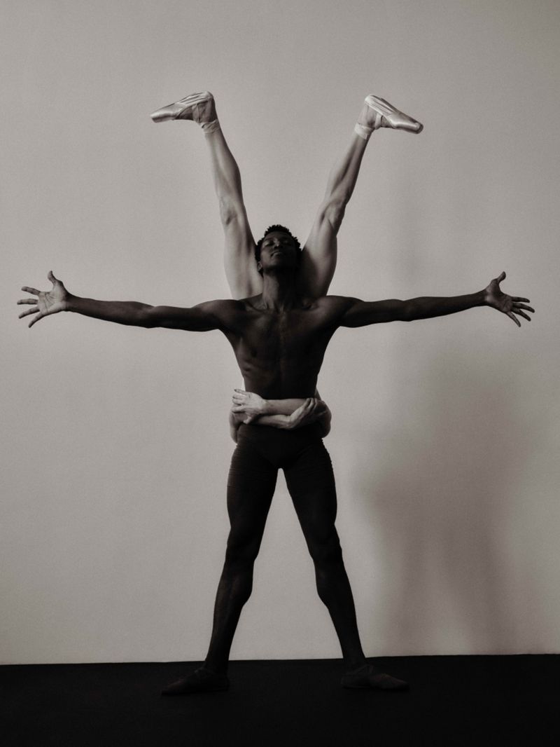 Two dancers in a black and white photo, one African American man supporting a Caucasian dancer in pointe shoes with arms outstretched.