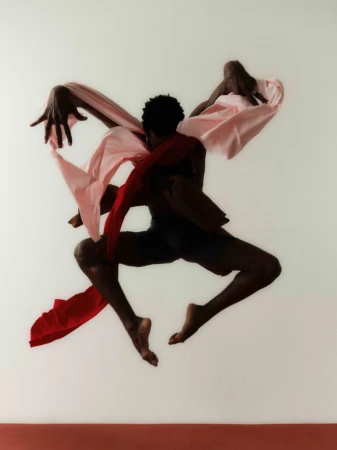 An athletic Black male dancer jumps against a white wall, draped with pink and red fabric, showcasing movement and expressive form.
