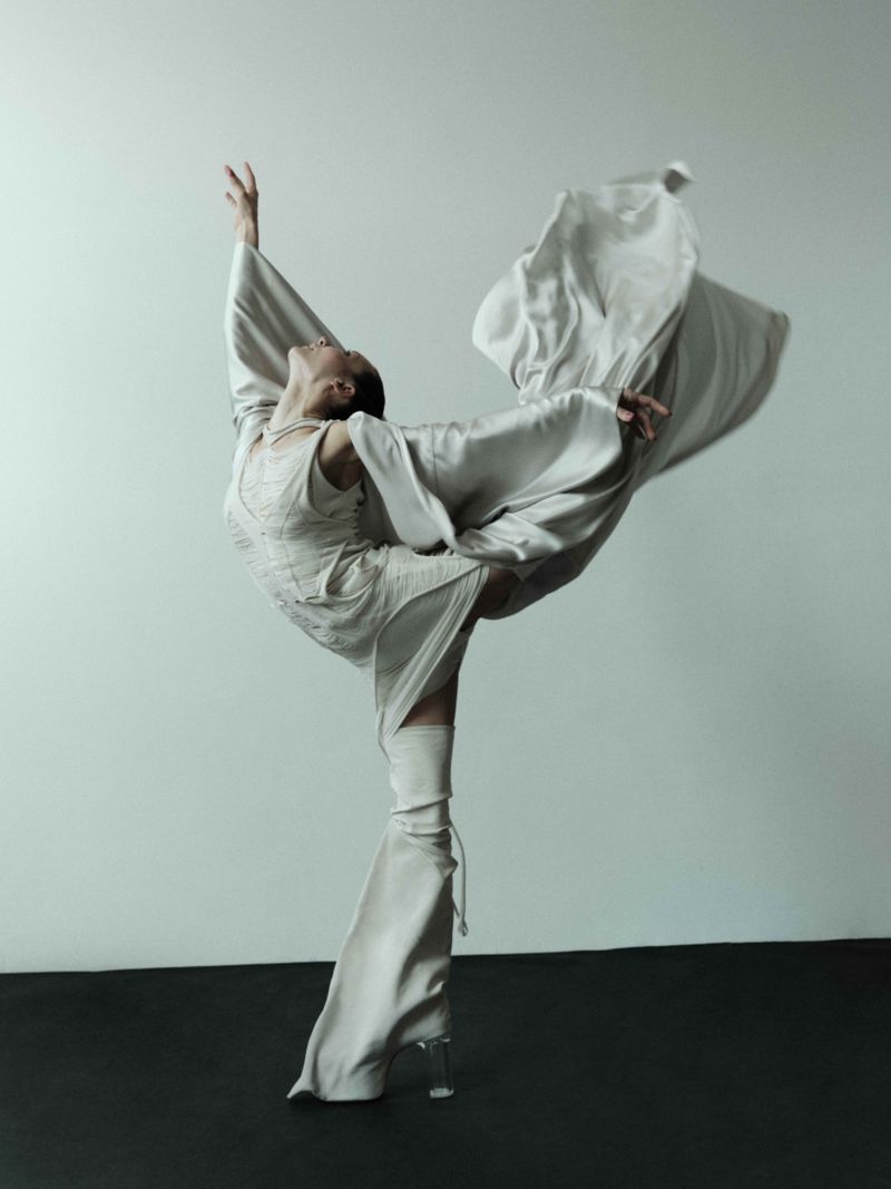 A solo adult female ballet dancer is posed in a complex full body gesture wearing a flowing neutral colored gown with clear heels indoors.