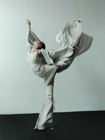 A solo adult female ballet dancer is posed in a complex full body gesture wearing a flowing neutral colored gown with clear heels indoors.