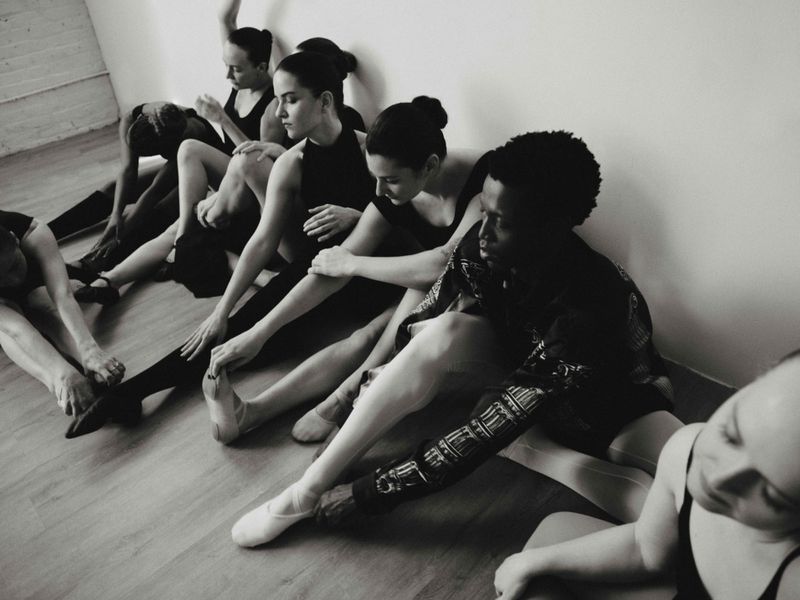 Monochromatic shot of a group of ballerinas in black leotards stretching on a wooden floor, leaning against a plain white wall.