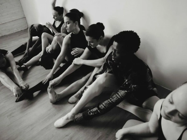 Monochromatic shot of a group of ballerinas in black leotards stretching on a wooden floor, leaning against a plain white wall.
