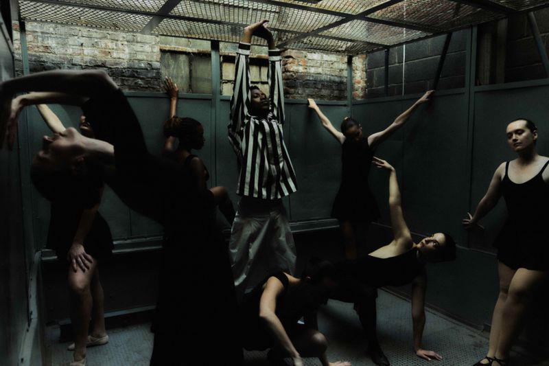 Group of dancers in an enclosed space with metal grates; one dancer stands out in a striped top, set against a grey and brick background.