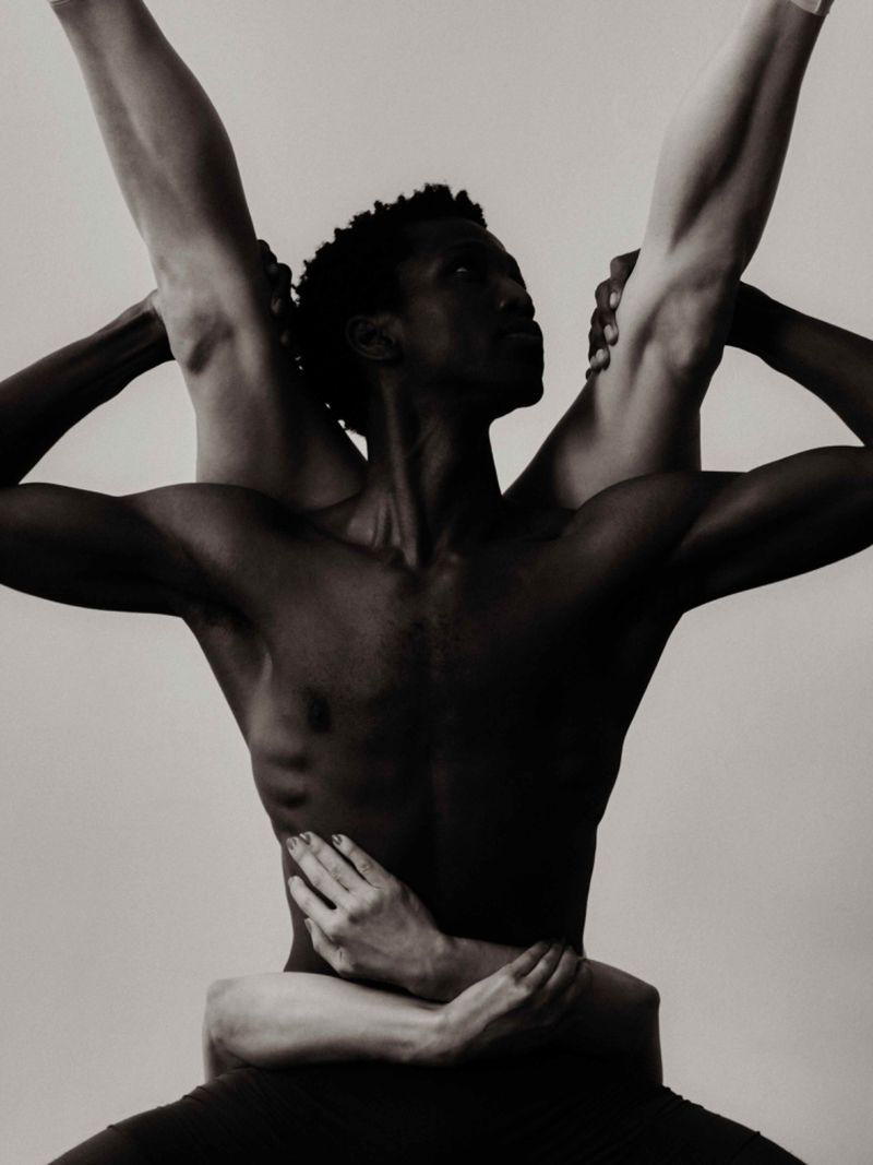 Monochromatic studio shot features an African American man's upper body, with arms and legs of a second, light-skinned person, holding him.