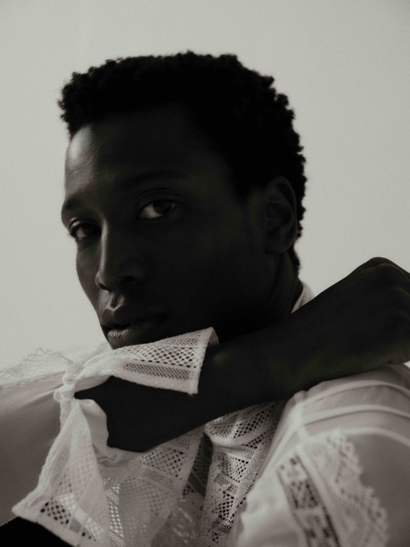 Portrait of a Black male model with short curly hair, posing in a white lace shirt against a bright backdrop. Fashion photography.
