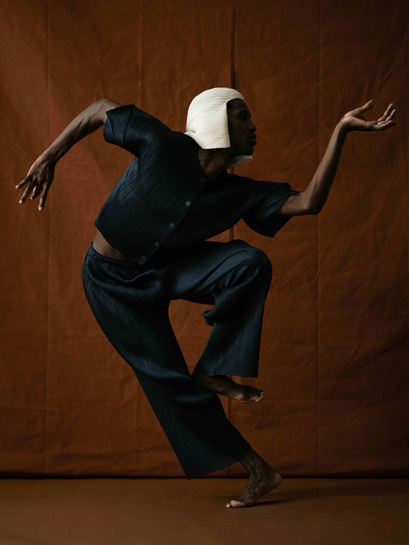 A black male model strikes a dynamic pose, wearing a dark, pleated outfit and a cream-colored hat against a terracotta background.