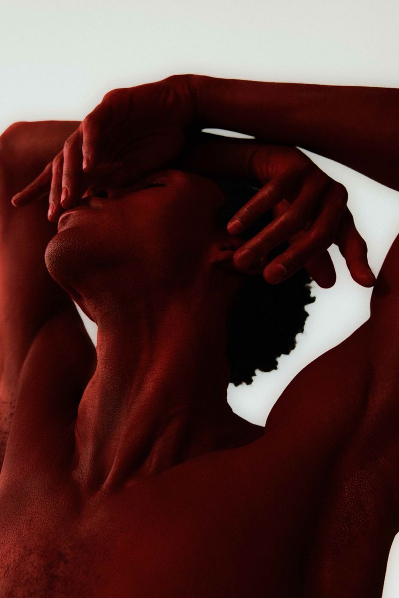 Portrait of a red-toned shirtless male with afro covering his face with his hands against a bright white background in a studio setting.