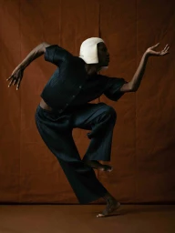 Dancer Calvin Royal III in dark pleated garments and white cap poses dynamically against a warm brown backdrop for Issue South America.