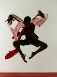 Black dancer in mid-air leap holding flowing red and pink fabric against a white background