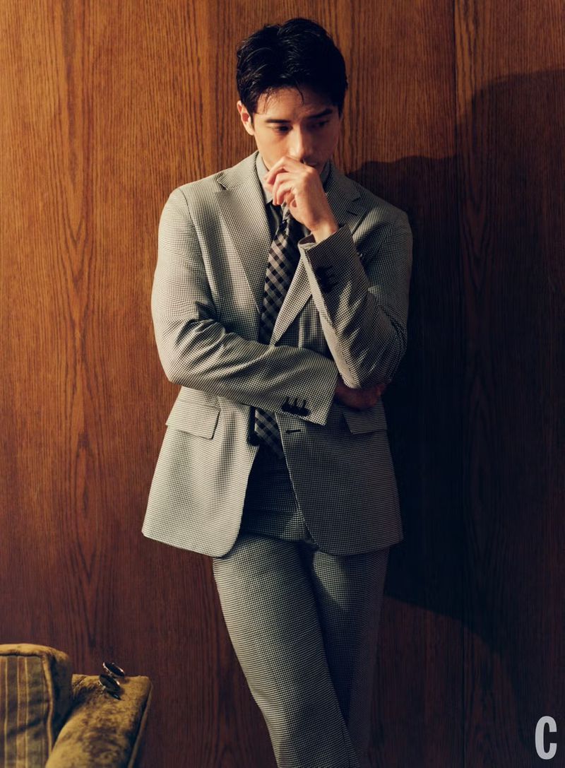 Portrait of an adult male in a grey suit and tie leaning against a paneled wall with his hand to his mouth in a dimly lit space.