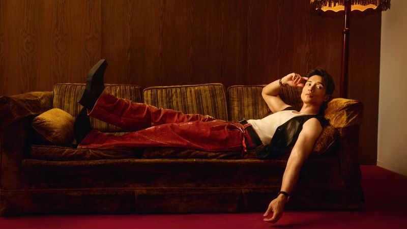 Man lies on a couch in a dimly lit room with wood paneling. He wears a white tank top, black vest, and red pants. Lamp stands nearby.
