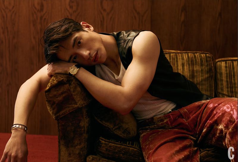 Handsome man with dark hair lounges on a plush brown couch wearing a black vest and red patterned pants in a dimly lit room.