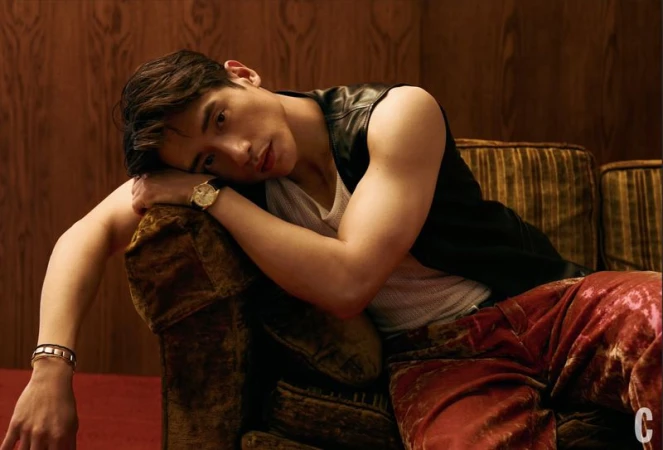 Handsome man with dark hair lounges on a plush brown couch wearing a black vest and red patterned pants in a dimly lit room.