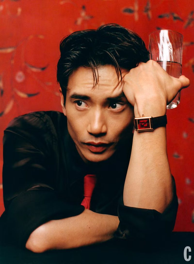 Portrait of a man in a black shirt holding a glass of water against a red patterned background, looking off to the side.
